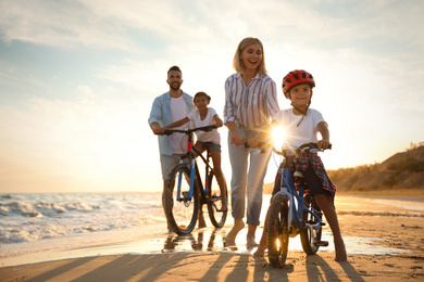 Happy parents teaching children to ride bicycles on sandy beach near sea at sunset