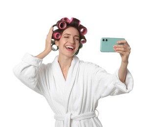 Happy young woman in bathrobe with hair curlers taking selfie on white background