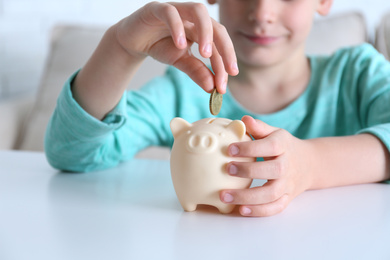 Little boy putting coin into piggy bank at white table indoors, closeup