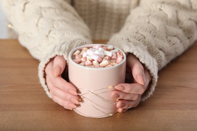 Woman in sweater holding cup of aromatic cacao with marshmallows on wooden table, closeup