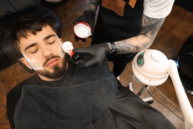 Professional hairdresser applying shaving foam onto client's beard in barbershop, closeup