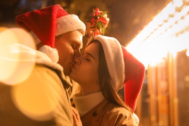 Happy couple in Santa hats kissing under mistletoe bunch outdoors, bokeh effect
