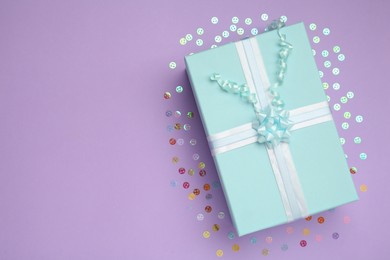 Light blue gift box and confetti on lilac background, flat lay. Space for text