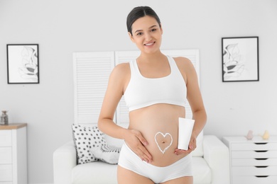 Pregnant woman with heart on her belly holding body cream at home