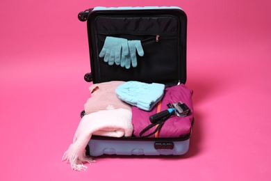 Open suitcase with warm clothes and action camera on color background. Winter vacation
