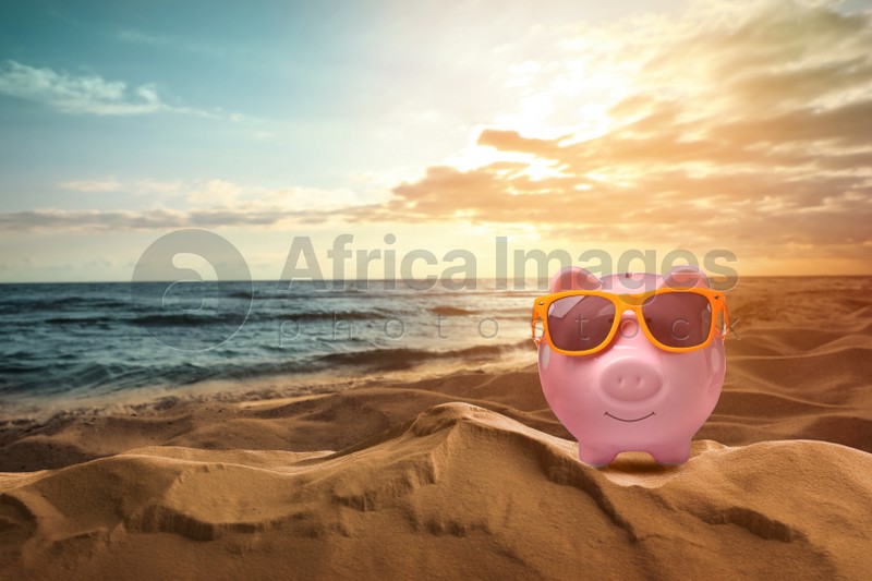 Saving money for summer vacation. Piggy bank with sunglasses on sandy beach near sea at sunset, space for text