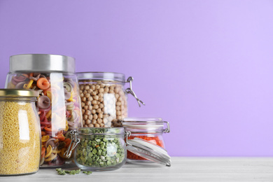 Jars with different cereals on white wooden table against violet background. Space for text