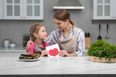 Photo of Little daughter congratulating mom with greeting card in kitchen. Happy Mother's Day