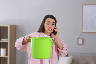 Young woman calling roof repair service while collecting leaking water from ceiling at home