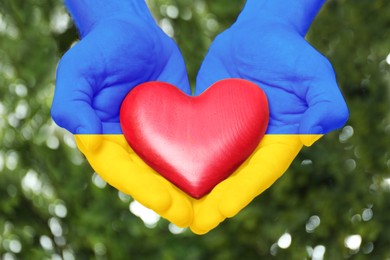 Man with hands in colors of Ukrainian flag holding red heart outdoors, closeup. Volunteering during war