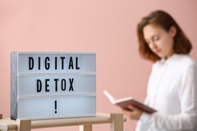 Woman reading book against pink background, focus on lightbox with phrase DIGITAL DETOX