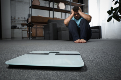 Scales and depressed overweight man on floor at home