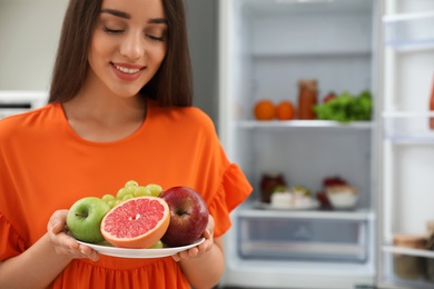 Concept of choice between healthy and junk food. Woman holding plate with fruits near refrigerator in kitchen