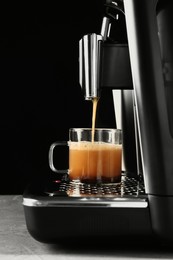 Modern espresso machine pouring coffee into glass cup on grey table against black background