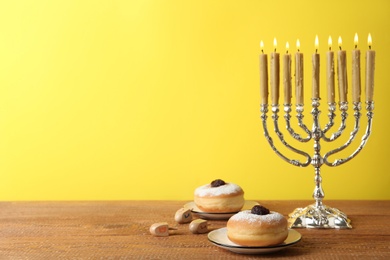 Silver menorah, dreidels with He, Pe, Nun, Gimel letters and sufganiyot on wooden table against yellow background, space for text. Hanukkah symbols