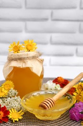 Delicious honey with dipper and different flowers on wicker mat against white brick wall