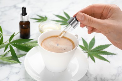 Woman dripping THC tincture or CBD oil into cup of coffee at white marble table, closeup