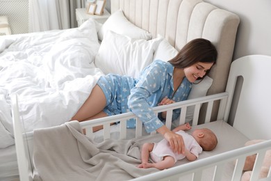 Happy young mother near bedside crib with her cute newborn baby at home