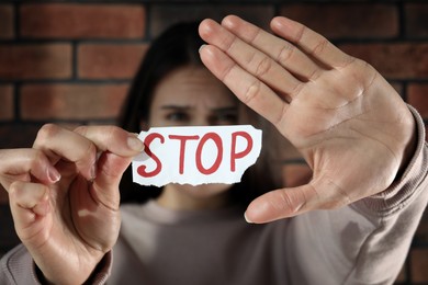 Photo of Domestic violence concept. Woman holding paper with written word Stop near brick wall, focus on hands