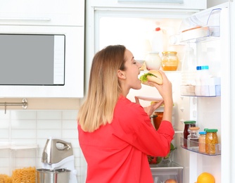 Photo of Hungry young woman eating sandwich near open refrigerator in kitchen. Failed diet