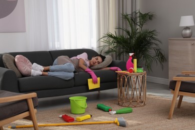 Tired young woman sleeping on sofa and cleaning supplies in living room