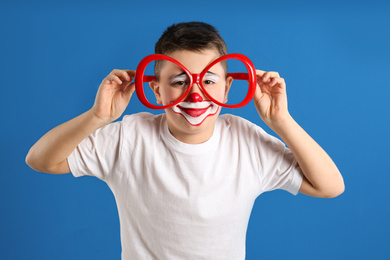 Preteen boy with clown makeup and party glasses on blue background. April fool's day