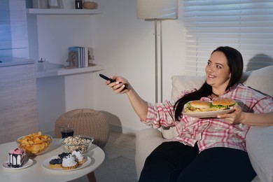 Happy overweight woman with unhealthy food watching TV on sofa at home
