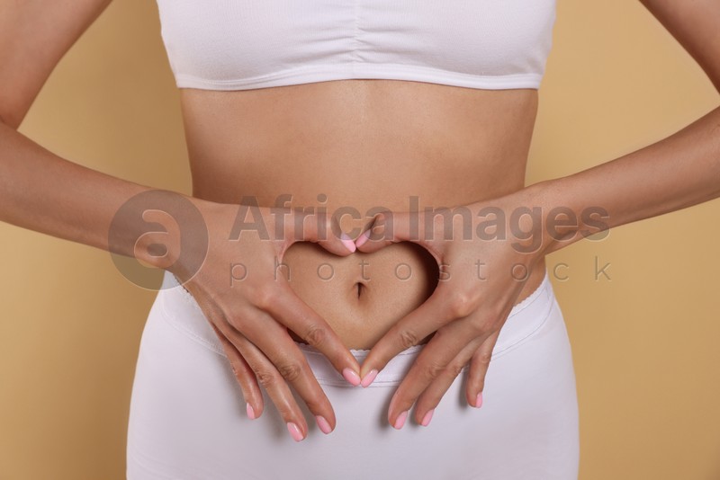 Woman in underwear making heart with hands on her belly against beige background, closeup. Healthy stomach