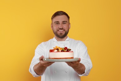 Happy professional confectioner in uniform holding delicious cake on yellow background