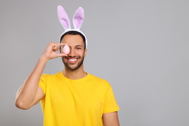 Photo of Happy African American man in bunny ears headband covering eye with Easter egg on gray background. Space for text