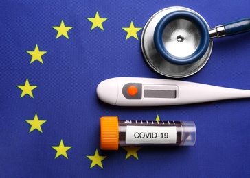 Test tube with blood sample, thermometer and stethoscope on European Union flag background, flat lay. Coronavirus outbreak