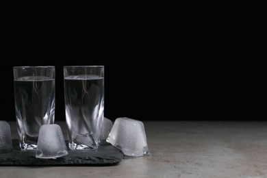 Shot glasses with vodka and ice cubes on grey tale against black background, space for text