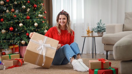 Photo of Young woman with beautifully wrapped gift near Christmas tree at home