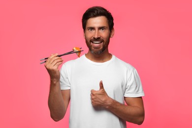 Happy man holding tasty sushi with chopsticks and showing thumbs up on pink background