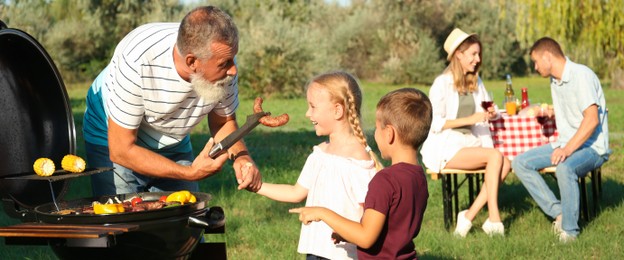 Grandfather with little kids cooking food on barbecue grill and their family in park. Banner design