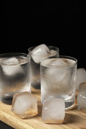 Shot glasses of vodka with ice cubes on wooden board against black background, closeup