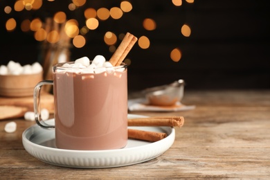 Glass cup of hot cocoa with aromatic cinnamon and marshmallows on wooden table against blurred lights. Space for text