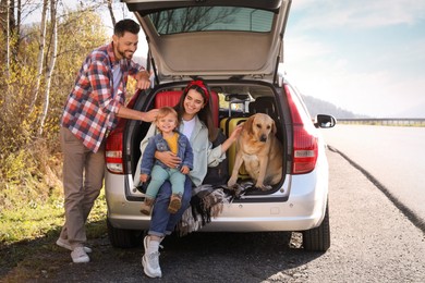 Photo of Parents, their daughter and dog sitting in car trunk near road. Family traveling with pet