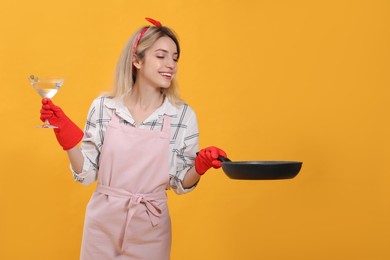 Young housewife with frying pan and glass of martini on yellow background. Space for text