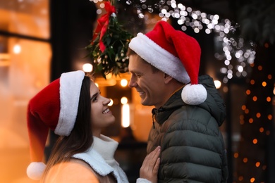 Happy couple in Santa hats standing under mistletoe bunch outdoors