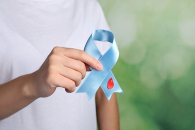 Woman holding light blue ribbon with paper blood drop against blurred green background, closeup. World Diabetes Day