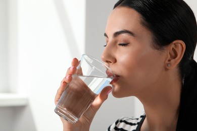 Beautiful young woman drinking water indoors, closeup. Refreshing drink