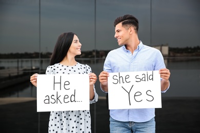 Lovely couple holding posters with text HE ASKED... SHE SAID YES after engagement outdoors