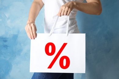 Woman holding paper shopping bag with percent sign near light blue wall, closeup. Discount concept