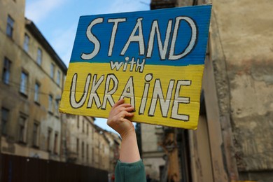 Photo of Young woman holding poster in colors of national flag and words Stand with Ukraine outdoors, closeup