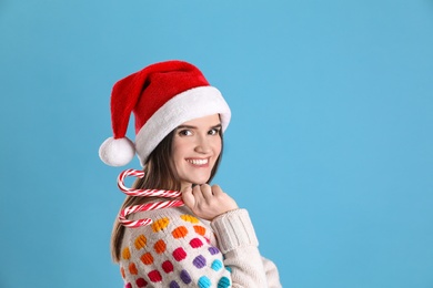 Pretty woman in Santa hat and festive sweater holding candy canes on light blue background, space for text