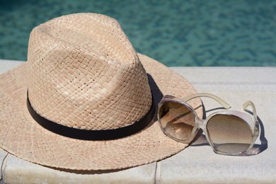 Stylish hat and sunglasses near outdoor swimming pool on sunny day, closeup. Beach accessories