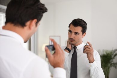Photo of Handsome young man using perfume near mirror indoors