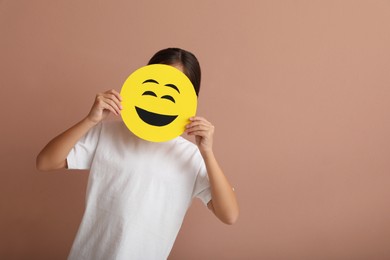 Little girl covering face with laughing emoji on pale pink background, space for text