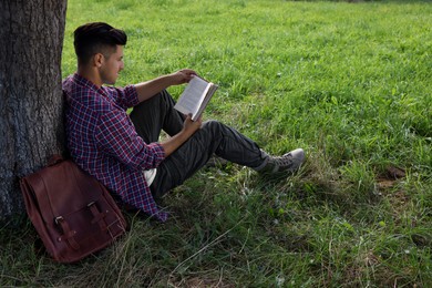 Handsome man reading book under tree on green meadow